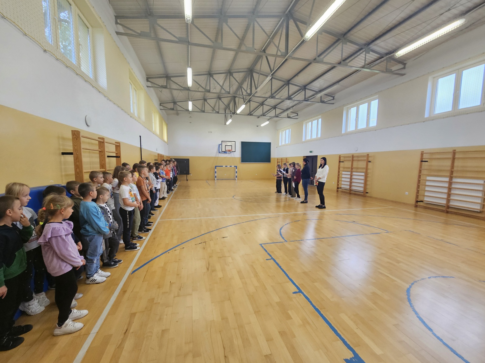 Uczniowie ustawieni w szeregu w sali gimnastycznej, obok stoją nauczyciele.